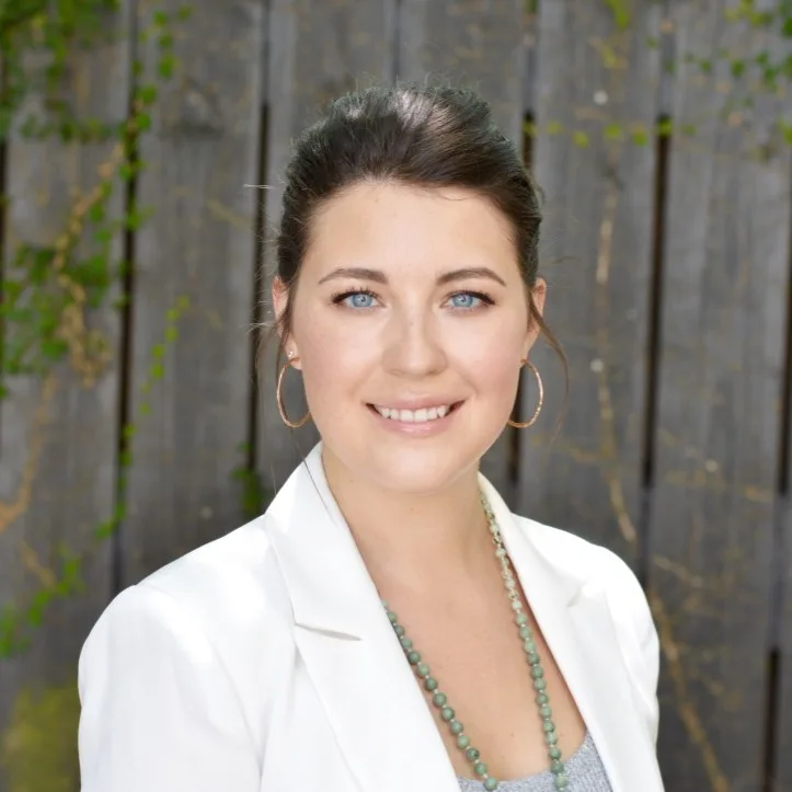 Portrait of a smiling woman with blue eyes in a white blazer, hoop earrings, and turquoise bead necklace, outdoors by a wooden fence.
