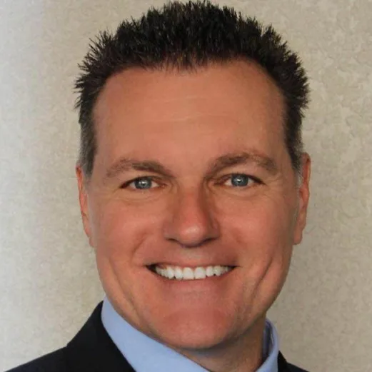 Head-and-shoulders portrait of a smiling man in a dark suit wearing a light blue shirt, against a neutral background.