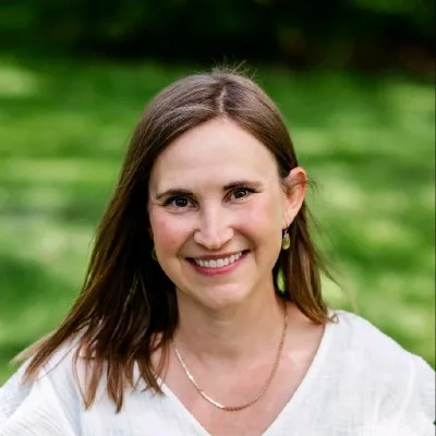 Smiling woman with shoulder-length brown hair in a white blouse, outdoors with a green blurred background.